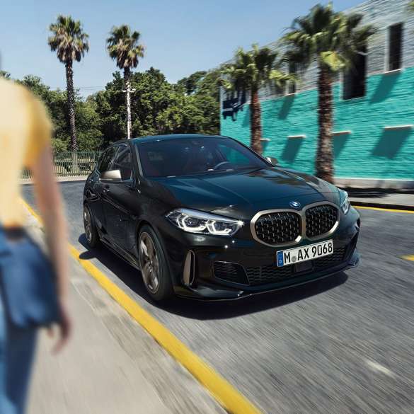 BMW M135i xDrive, three-quarter front view F40. BMW M135i xDrive in Black Sapphire metallic, three-quarter front view driving, with woman in foreground F40.