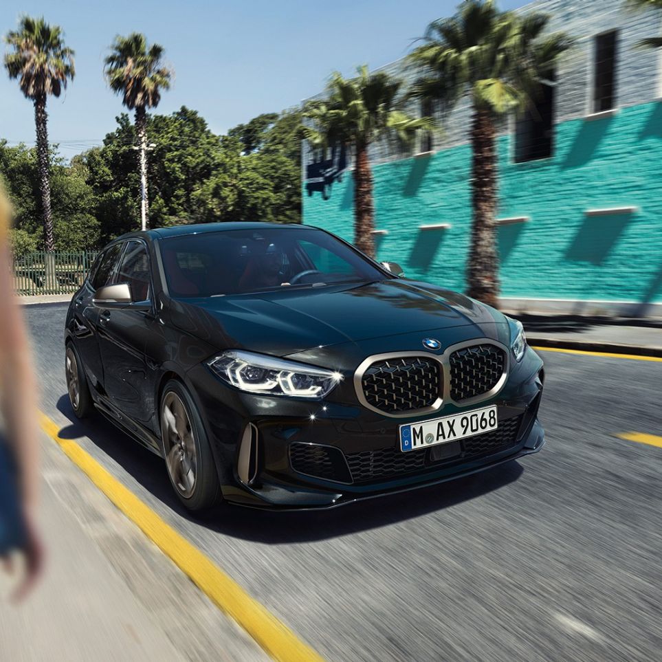 BMW M135i xDrive, three-quarter front view F40. BMW M135i xDrive in Black Sapphire metallic, three-quarter front view driving, with woman in foreground F40.