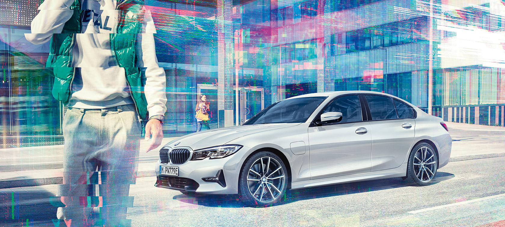 BMW 3 Series Sedan, side shot BMW 330e G20. Three-quarter side shot of the standing BMW 330e Sedan in front of an urban glass building with walking passengers G20.
