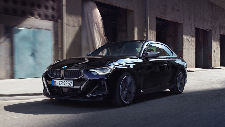 BMW M240i xDrive Coupé G42 2021 Black Sapphire metallic three-quarter side view driving in front of warehouses