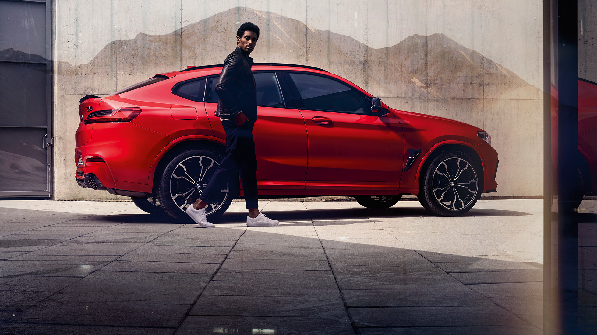 BMW X4 M Competition SUV, side view with young man F98. BMW X4 M Competition SUV in Toronto Red metallic, exterior, side view with young man F98.